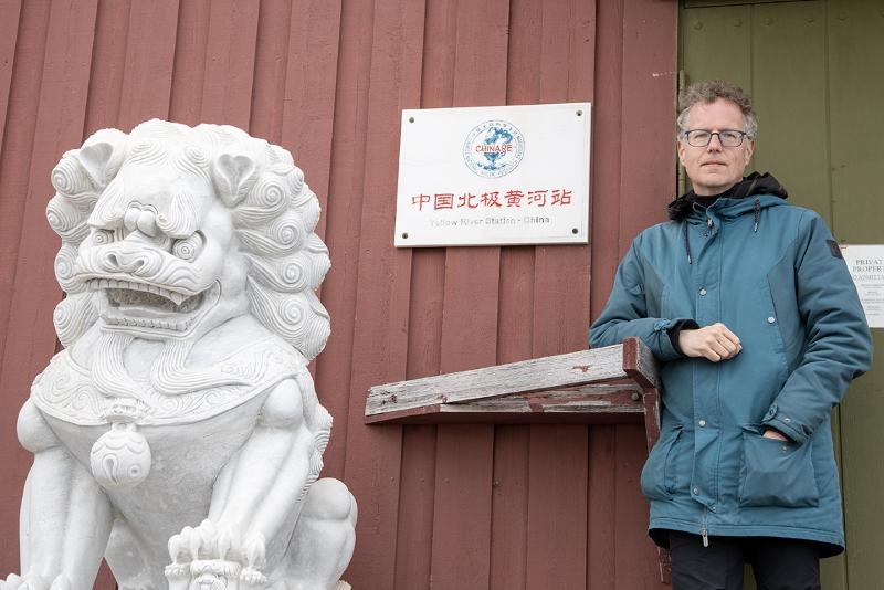 marc lanteigne yellow river station ny-ålesund foto karine aarskog uit.jpg Ein mann henslengt foran ein raud bygning. Ved sidan han ein dør, og ved sisan av den igjen ein statue av ein løve i kinesisk stil.